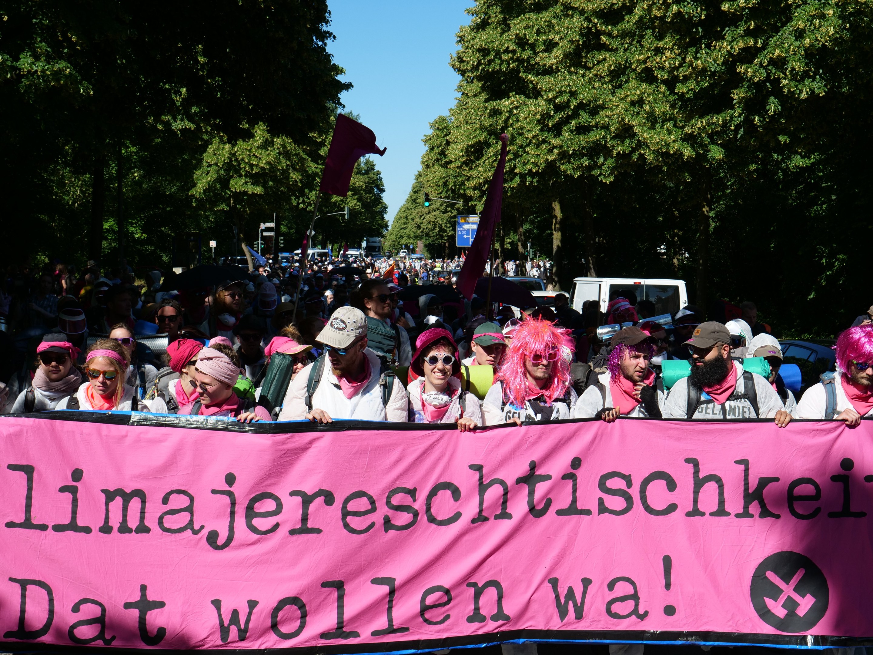 Eine Gruppe von Menschen marschiert auf einer Straße, hält ein pinkes Banner mit der Aufschrift "Women's March in Berlin" und trägt teilweise Mützen und Schutzbrillen, während andere Fahnen halten; Bäume, Fahrzeuge und Himmel sind im Hintergrund zu sehen.