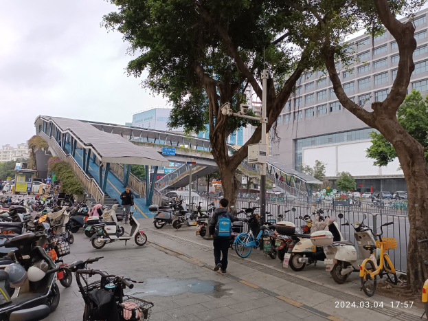 Eine belebte Straße mit parkenden E-Scootern, Fussgängern auf dem Gehweg, Bäumen, Gebäuden und bewölktem Himmel mit einem Wasserzeichen.