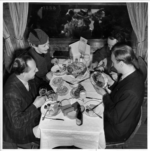 Eine Gruppe von Menschen sitzt an einem Tisch mit Tellern, Gläsern, Flaschen und Besteck, mit Vorhängen im Hintergrund.