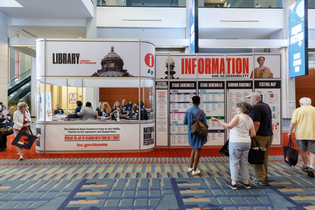 Eine Gruppe von Menschen auf dem Nationalen Buchfest, einige sitzen und einige stehen, vor einem Stand mit Bannern, einem Anzeigebildschirm, einer Treppe, einem Geländer und einem Dach mit Deckenleuchten.