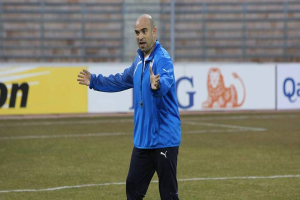 Ein Mann in einer blauen Jacke steht auf einem Fußballfeld, umgeben von Schildern mit Text und Logos, entschlossen.