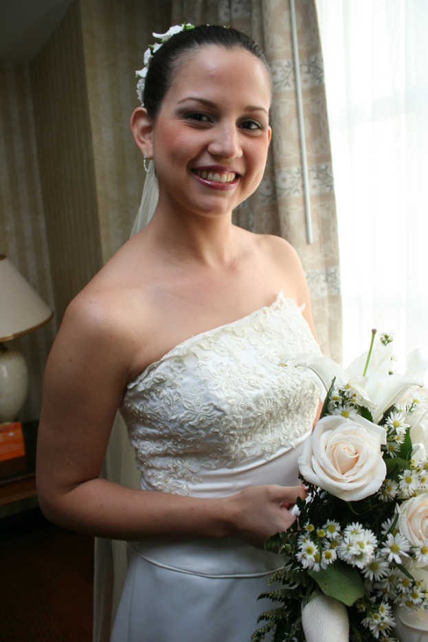 Eine Braut steht lachend, eine Blume haltend, vor einem Vorhang und einer Wand und eine Lampe auf einem Tisch links.