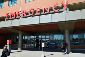 Eine Gruppe von Menschen vor einem Notfallgebäude mit Glaswänden, Säulen, einem Namensschild, Schildern auf Stangen und einem Mülleimer.