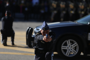 Ein Polizist in Uniform kümmert sich auf der Straße um einen Mann, mit einem Auto und einer Person im Hintergrund.