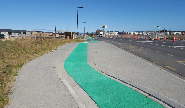 Eine Straße mit einem grünen Radweg an der Seite, Gras auf der linken Seite und Verkehrskegel auf der rechten Seite, mit Laternenmasten, Bäumen, Gebäuden und einem klaren blauen Himmel im Hintergrund.