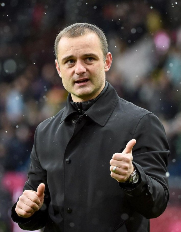 Ein Mann in schwarzer Jacke zeigt den Daumen hoch, trägt eine Uhr und hat einen unscharfen Hintergrund.