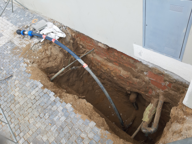 Ein großes Loch im Boden neben einem Gebäude mit verstreuten Rohren und Gegenständen, ein Metallobjekt links und eine Metallbox an der rechten Wand, was auf eine laufende Kanalsanierung hinweist.