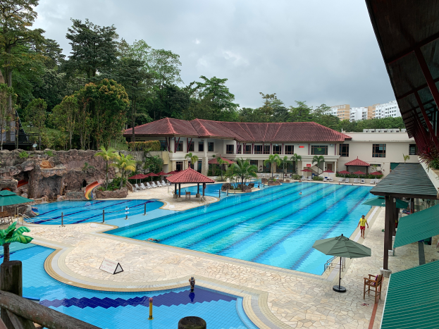 Ein großer Swimmingpool im Ritz-Carlton, Kota Kinabalu, voller Menschen, umgeben von Schirmen, Stühlen, Tischen, Pflanzen, Bäumen, Felsen und Gebäuden, mit einem Schuppen auf der rechten Seite und dem Himmel im Hintergrund.
