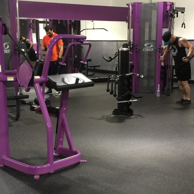 Eine Person in einem schwarzen Kleid, einer Mütze und Schuhen steht neben einer anderen Person in einem orangen T-Shirt und Schuhen in einem Fitnessstudio mit Geräten und einem Spiegel im Hintergrund.