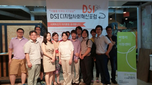 Eine Gruppe von Menschen, die in einem Raum stehen und lachen, mit einem Banner im Hintergrund und Deckenlampen auf der rechten Seite.