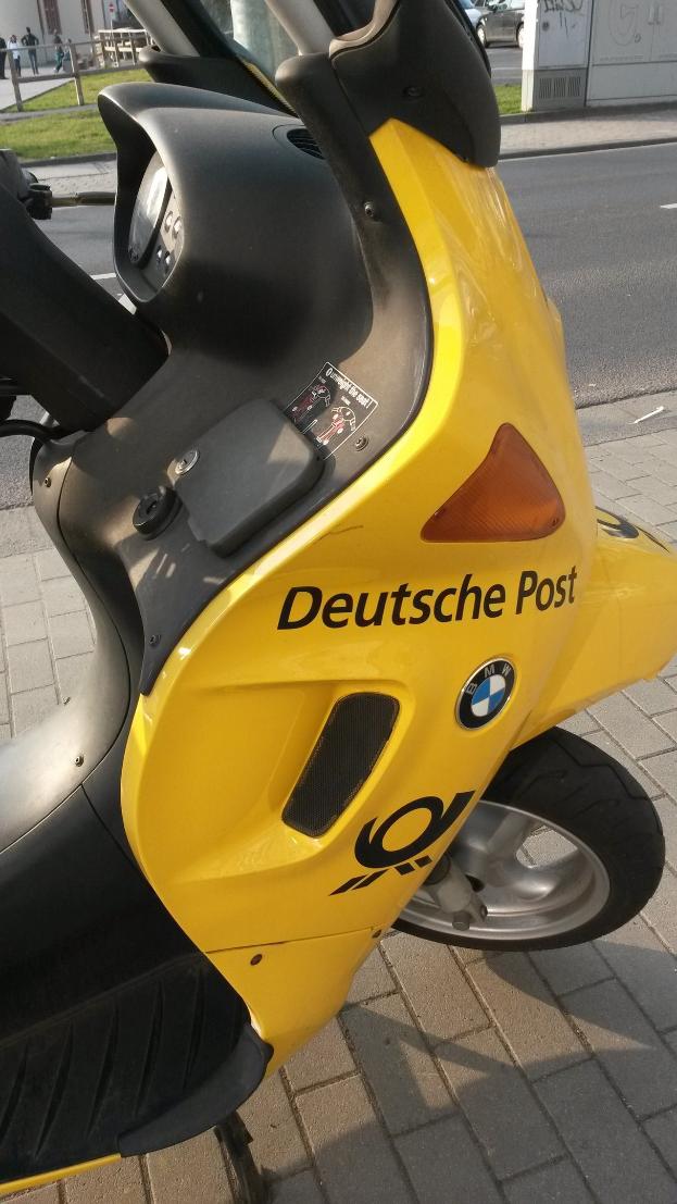 Ein gelber Scooter steht am Straßenrand mit einem sichtbaren Kennzeichen, umgeben von Gebäuden, Bäumen, Polen, Brettern und anderen Fahrzeugen auf der Straße.