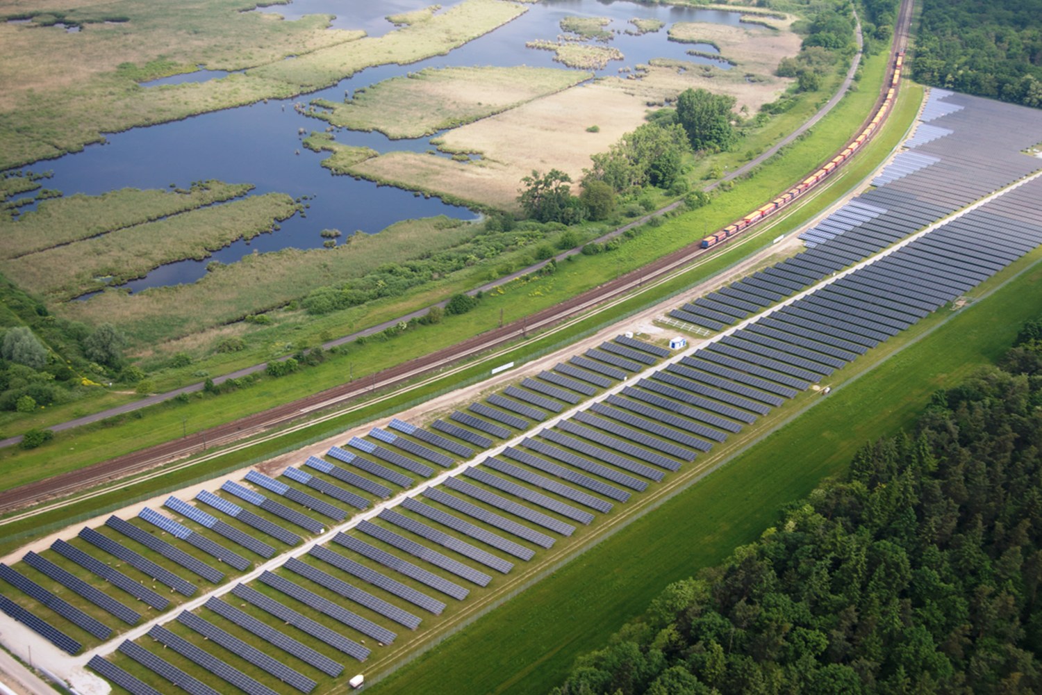 Luftaufnahme eines Solarparks mit Panelen in einem Feld, umgeben von Bäumen, Gras, Pflanzen und Wasser, mit einem Zug auf einer nahen Bahnlinie.
