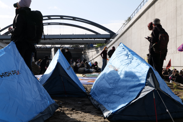 Eine Gruppe von Menschen sitzt auf einem sandigen Strand in der Nähe von Zelten, mit einer Wand und einer Brücke im Hintergrund, die an einer Klimawandel-Demonstration teilnehmen, unter einem bewölkten Himmel.
