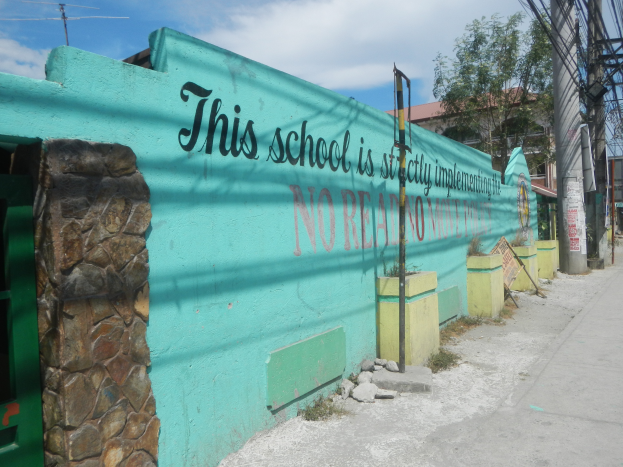 Eine grüne Wand mit Graffiti, das 'Diese Schule ist keine Stadt' lautet, umgeben von Gebäuden, Bäumen, Pfählen, Drähten, Brettern und anderen Gegenständen, unter einem bewölkten Himmel.