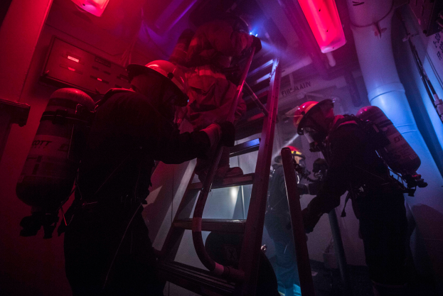 Feuerwehrleute mit Helmen und Zylindern klettern eine Leiter in einem dunklen, rot beleuchteten Raum mit einer Wand aus Rohren und Gegenständen auf der rechten Seite hoch.