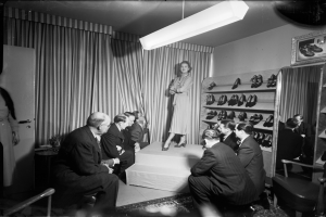 Eine Frau in einem Kleid steht auf einer Bühne vor sitzenden Menschen, mit Vorhängen im Hintergrund, Schuhen auf Gestellen und einem Rahmen an der Wand, alles in Schwarz-Weiß dargestellt.