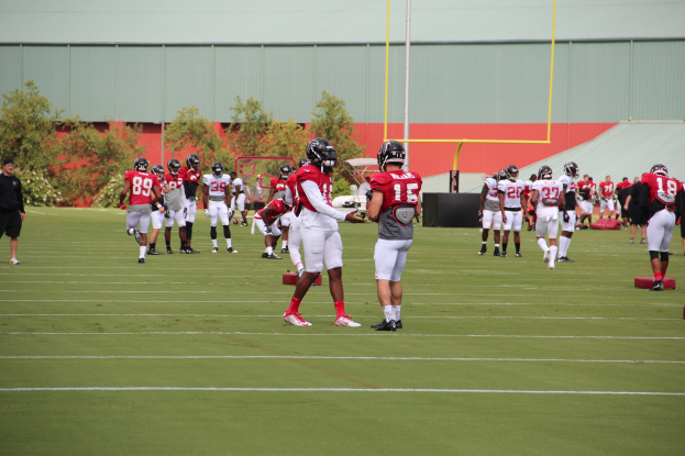 Gruppe von Menschen, die Fußball auf einem Rasenfeld mit Helmen und Stollenschuhen spielen, umgeben von Bäumen, Pfählen, einer Wand und einem klaren blauen Himmel.