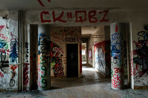 Ein verlassenes Gebäude mit Graffiti an den Wänden und Säulen, sichtbarer Boden, Rohre an den Wänden und eine Tür im Hintergrund, die den Verfall der Stadt zeigt.