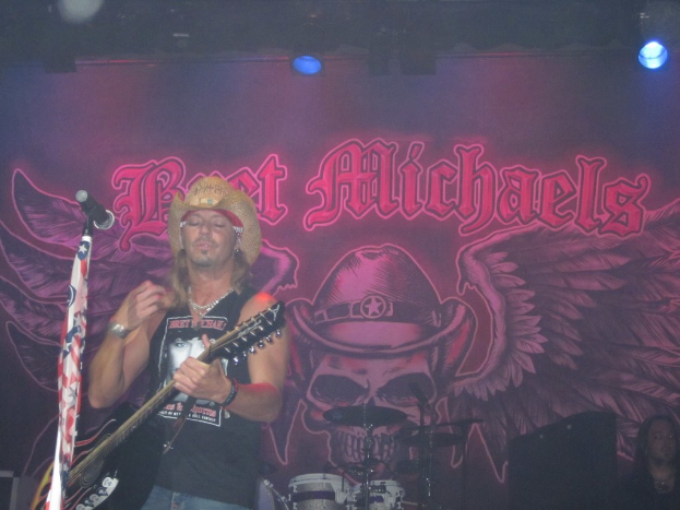 Eine Musik-Konzert-Szene mit einem Gitarristen auf der Bühne, einer Person in der unteren rechten Ecke, einem Banner im Hintergrund und Drogen in der Mitte unten, mit Scheinwerfern oben.