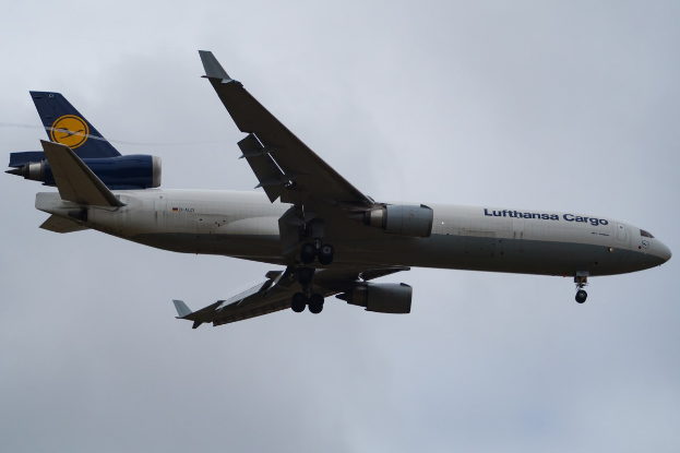 Lufthansa Cargo Boeing 777-300ER im Flug mit sichtbarem Text auf dem Rumpf, ausgefahrenen Tragflächen, aktiven Triebwerken, vor einem Himmel mit weißen Wolken.