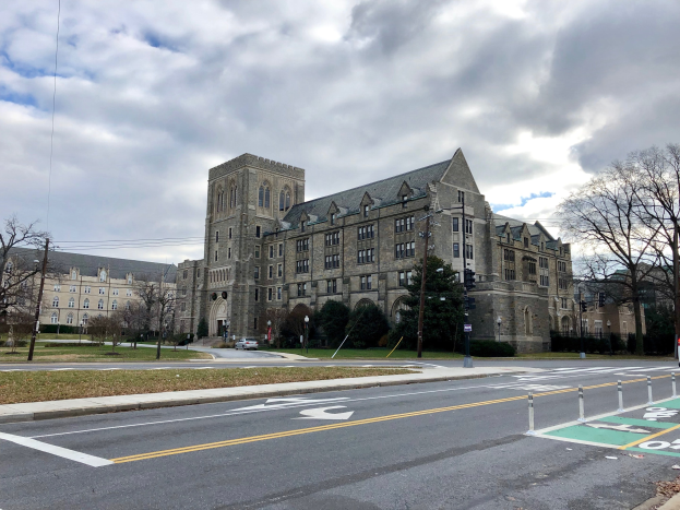 Großes Gebäude mit vielen Fenstern, als Universität von Pennsylvania bezeichnet, umgeben von Bäumen, Laternen, Ampeln und Fahrzeugen an einem bewölkten Tag.