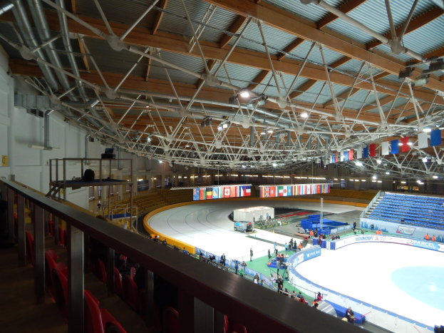 Große Indoor-Eisbahn voller Menschen, umgeben von Sitzplätzen, Geländern, Brettern, Beleuchtungskörpern und strukturellen Elementen, mit einer Wand im Hintergrund.