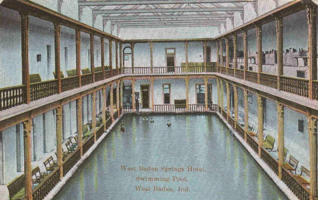 Eine alte Postkarte, die ein Hallenbad im West Baden Springs Hotel zeigt, mit Menschen, die schwimmen, und verschiedenen architektonischen Elementen drumherum.