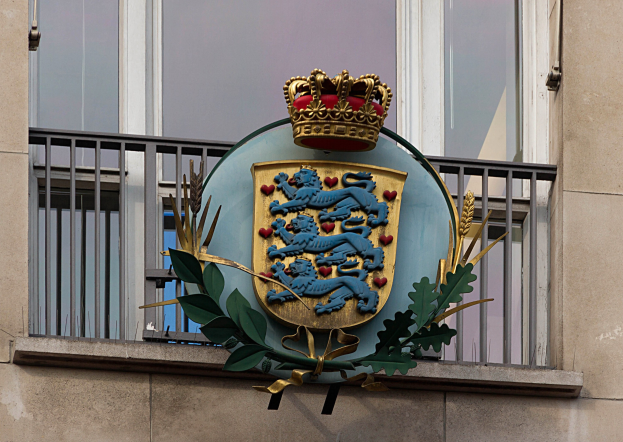 Das britische Königswappen auf der Außenseite eines Gebäudes, begleitet von einem Logo mit Krone und Blättern, mit Fenstern und Metallgittern im Hintergrund.