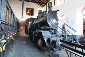 Eine alte Dampflokomotive in einem Museum ausgestellt, umgeben von Geländern, Treppen, Informationsschildern und anderen Ausstellungsstücken.