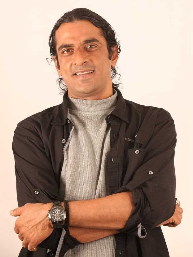 Ein Mann in einem schwarzen und aschfarbenen Kleid mit verschr├Ąnkten Armen, der eine Uhr tr├Ągt, vor einem wei├čen Hintergrund steht und nachdenklich wirkt.