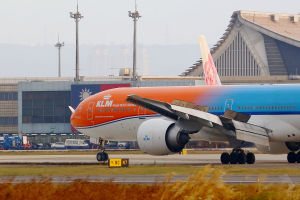 Ein KLM Boeing 777-300ER Flugzeuge ist auf der Rollbahn am Frankfurter Flughafen positioniert, mit Gras und Pflanzen am Boden sichtbar. Im Hintergrund gibt es Gebäude, Fahrzeuge, Masten und einen klaren blauen Himmel.