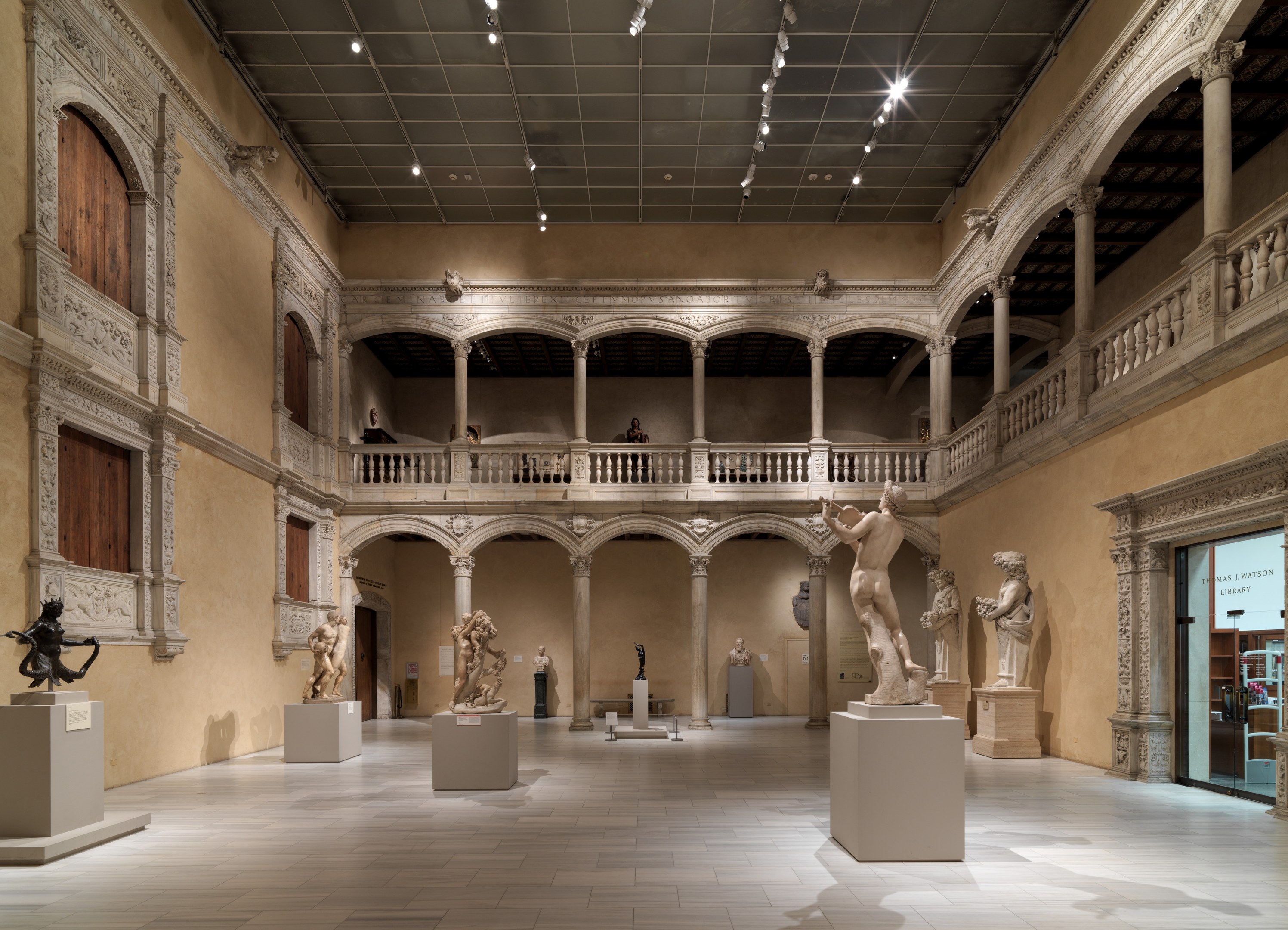 Ein geräumiger Museumsgalerie mit zahlreichen Statuen und Skulpturen auf Sockeln, mit architektonischen Elementen wie Wänden, Säulen, Geländern und Deckenleuchten und einigen Besuchern.