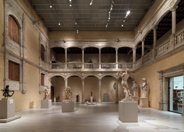 Ein geräumiger Museumsgalerie mit zahlreichen Statuen und Skulpturen auf Sockeln, mit architektonischen Elementen wie Wänden, Säulen, Geländern und Deckenleuchten und einigen Besuchern.
