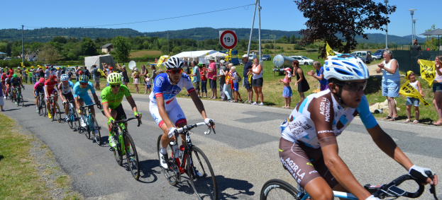 Eine Gruppe von Radfahrern fährt eine Straße entlang, während Zuschauer am Straßenrand stehen, einige tragen Helme und Schutzbrillen, andere halten Fahnen, mit Bäumen, Fahrzeugen, Zelten, Gebäuden und Hügeln im Hintergrund unter einem klaren blauen Himmel.