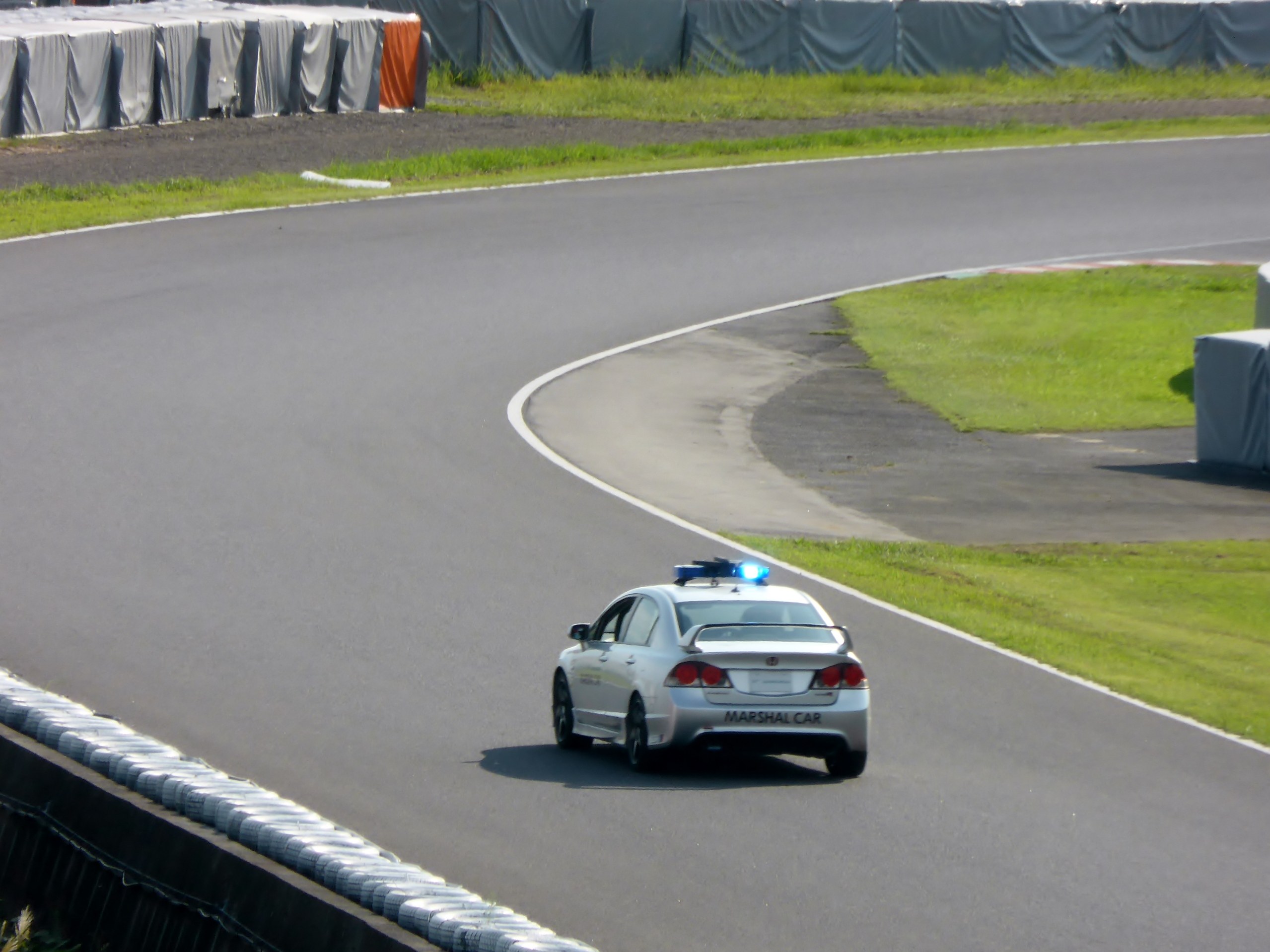 Ein Polizeiwagen auf einer Rennstrecke mit Gras auf beiden Seiten und einigen Menschen und Zelten im Hintergrund.