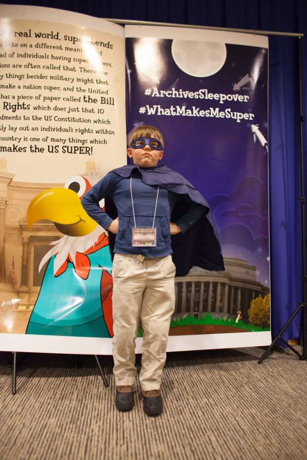 Ein junger Junge in einem Superheldenkostüm steht vor einem Banner mit Text und einem Vogelbild, mit einem Vorhanghintergrund, trägt Schuhe.