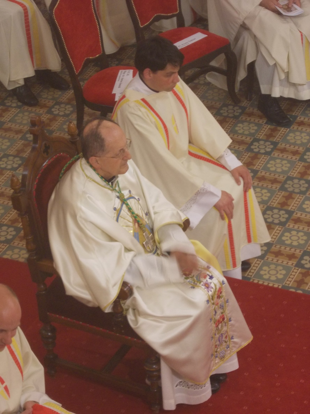 Eine Gruppe von Priestern in weißen Gewändern sitzt auf Stühlen auf einem roten Teppichboden in einer Kirche, einige halten Papiere.
