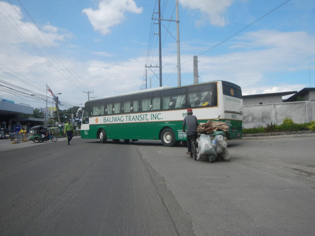 Ein grüner und weißer Bus fährt auf einer Straße mit Fußgängern, einem Radfahrer, Gebäuden, Bäumen und Strommasten im Hintergrund.
