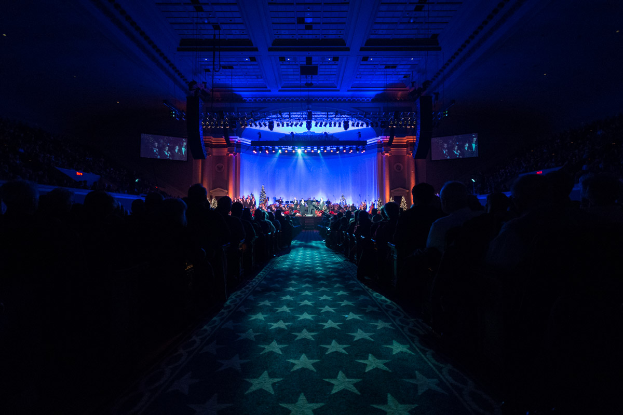 Ein großer Auditorium mit Sitzenden, eine Bühne mit Musikern und eine Decke mit Lichtern und Fernsehern, was auf einen Konzertsaal für die Nationale Convention 2019 hindeutet.