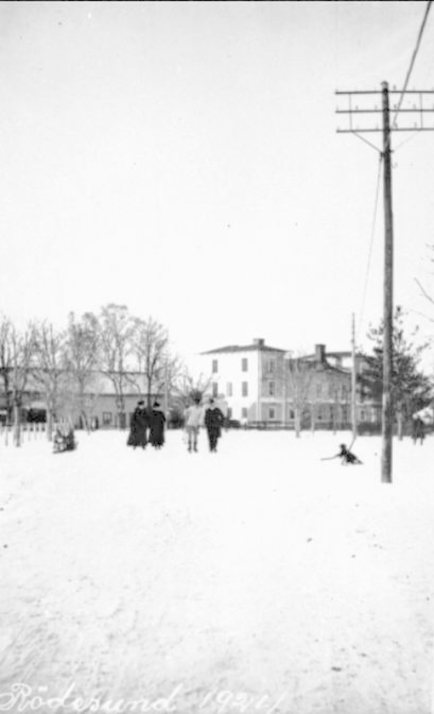 Eine Gruppe von Menschen geht auf einer schneebedeckten Straße in Rodesund, Deutschland im Jahr 1921 entlang, gesäumt von Bäumen, Gebäuden und Strommasten mit Drähten, unter einem sichtbaren Himmel, mit Text am unteren Rand des Bildes.