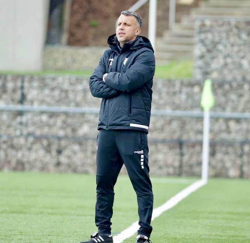 Ein Fußballtrainer steht auf einem Feld mit verschränkten Armen, trägt eine schwarze Jacke und Hose, vor einem Zaun, Treppe und Wand.