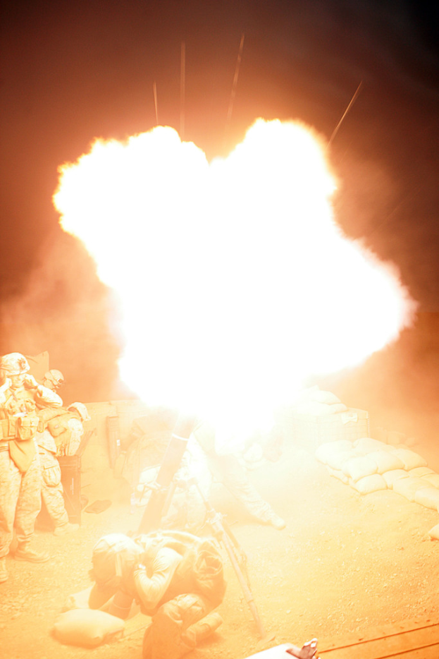 Mehrere Personen in Uniformen und Helmen auf einem Gelände mit ein paar Säcken drumherum, mit einem Feuer in der Mitte der Szene.