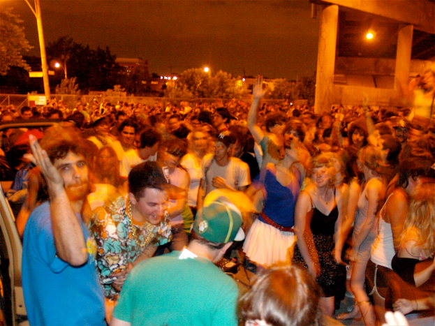 Menschen tanzen vorne vor einem Konzert mit Lichtern, Pfählen, Bäumen und anderen Gegenständen im Hintergrund.