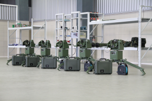 Gruppe von Militärrobotern auf einem Boden mit Koffern und Objekten auf Gestellen, vor einer Wand mit Geländer.