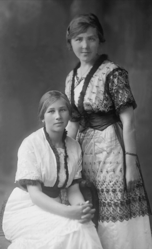 Zwei Frauen in Kleidern sitzen nebeneinander auf einer Bank, eine steht dahinter, vor leicht unscharfem Hintergrund mit Text unten.