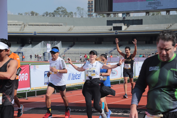 Gruppe von Menschen, die auf einer Außenbahn in einem Stadion laufen, mit Bannern, einer Anzeigetafel, einem Gebäude, Bäumen und einem bewölkten Himmel; einige Läufer tragen Turnschuhe und Hüte.