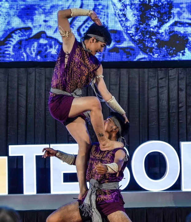 Zwei Akrobaten, ein Mann und eine Frau in purpurfarbenen Kleidern, führen eine Hebung auf der Bühne vor einem Publikum durch, mit einem schwarzen Vorhang und einer Leinwand im Hintergrund.