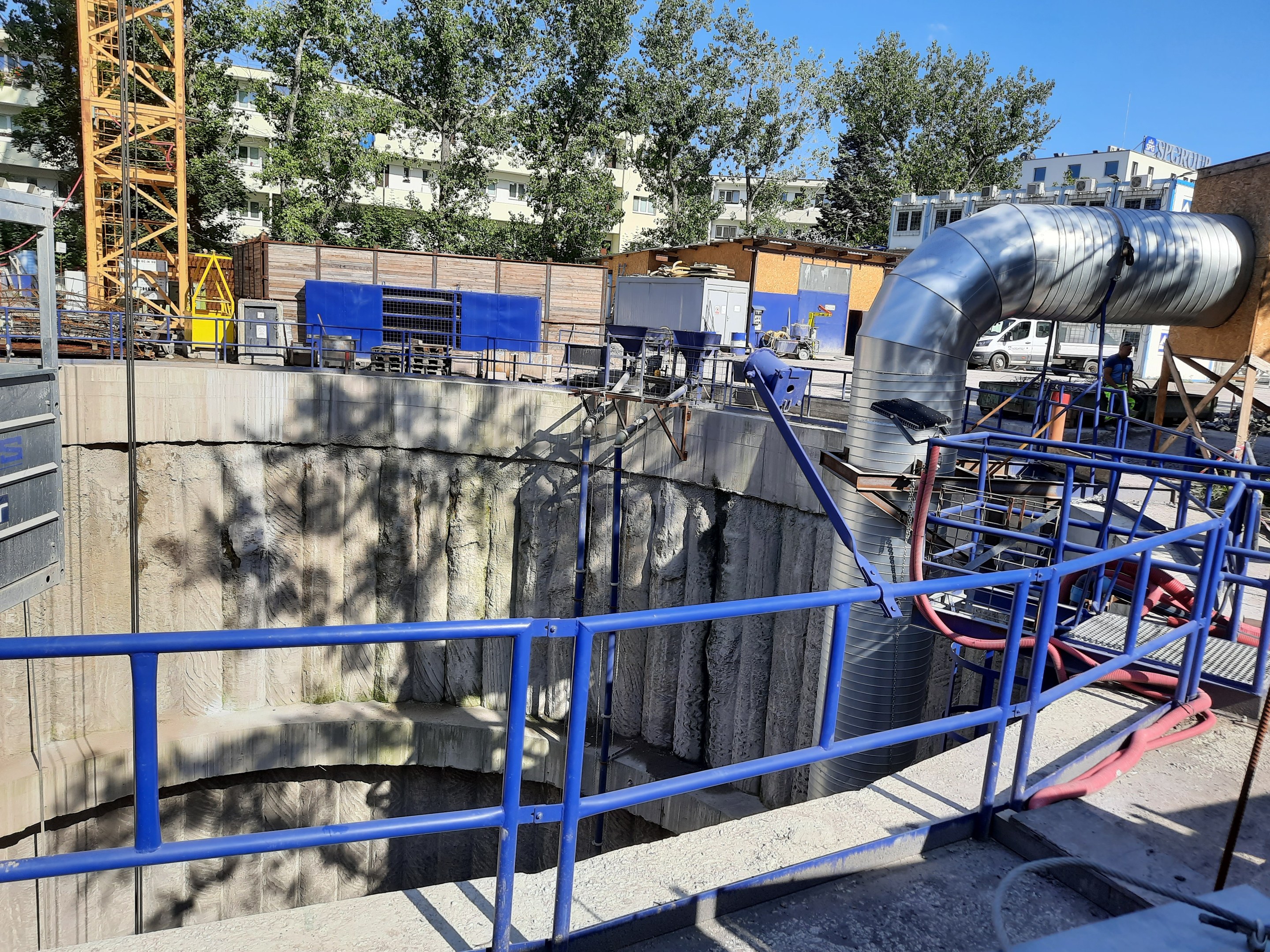 Große Metallrohr an einer Abwasserbehandlungsanlage-Baustelle, umgeben von Geländern, Rohren und Maschinen, mit Gebäuden, Bäumen und einem Turm im Hintergrund.