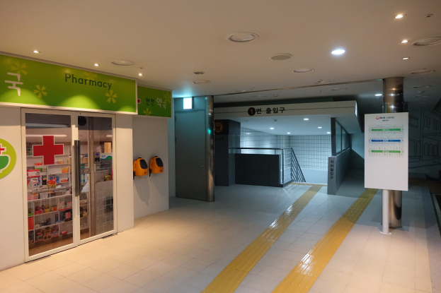 Apothekeneingang mit einem "Apotheke"-Schild links, einer Tafel mit Text rechts, beleuchteten Lampen oben und Hintergrundobjekten.