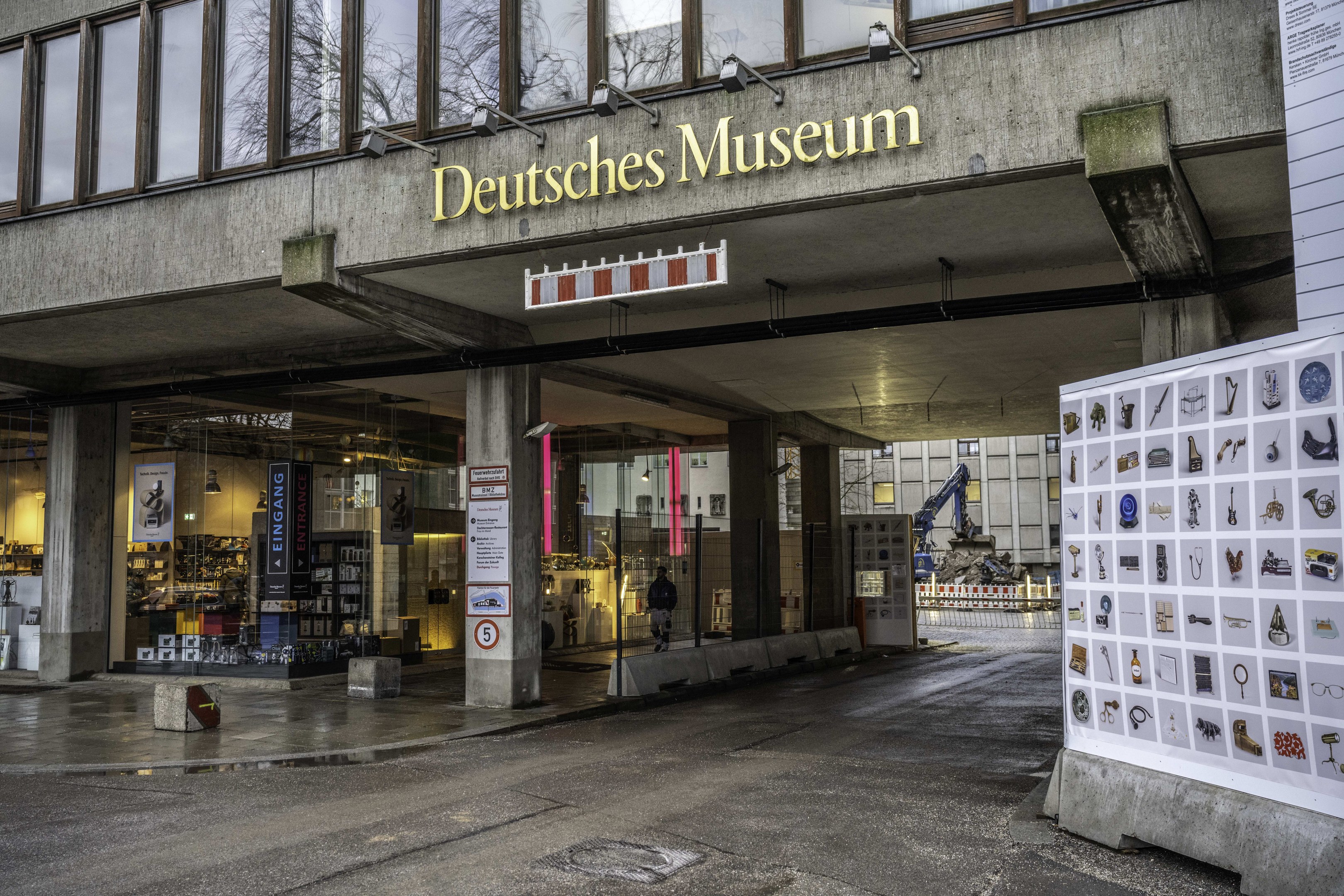 Außenansicht des Deutschen Museums in Berlin mit Glasfenstern, Säulen, einem Namensschild und einer Informationsanzeigetafel, umgeben von Bäumen und anderen Gebäuden.